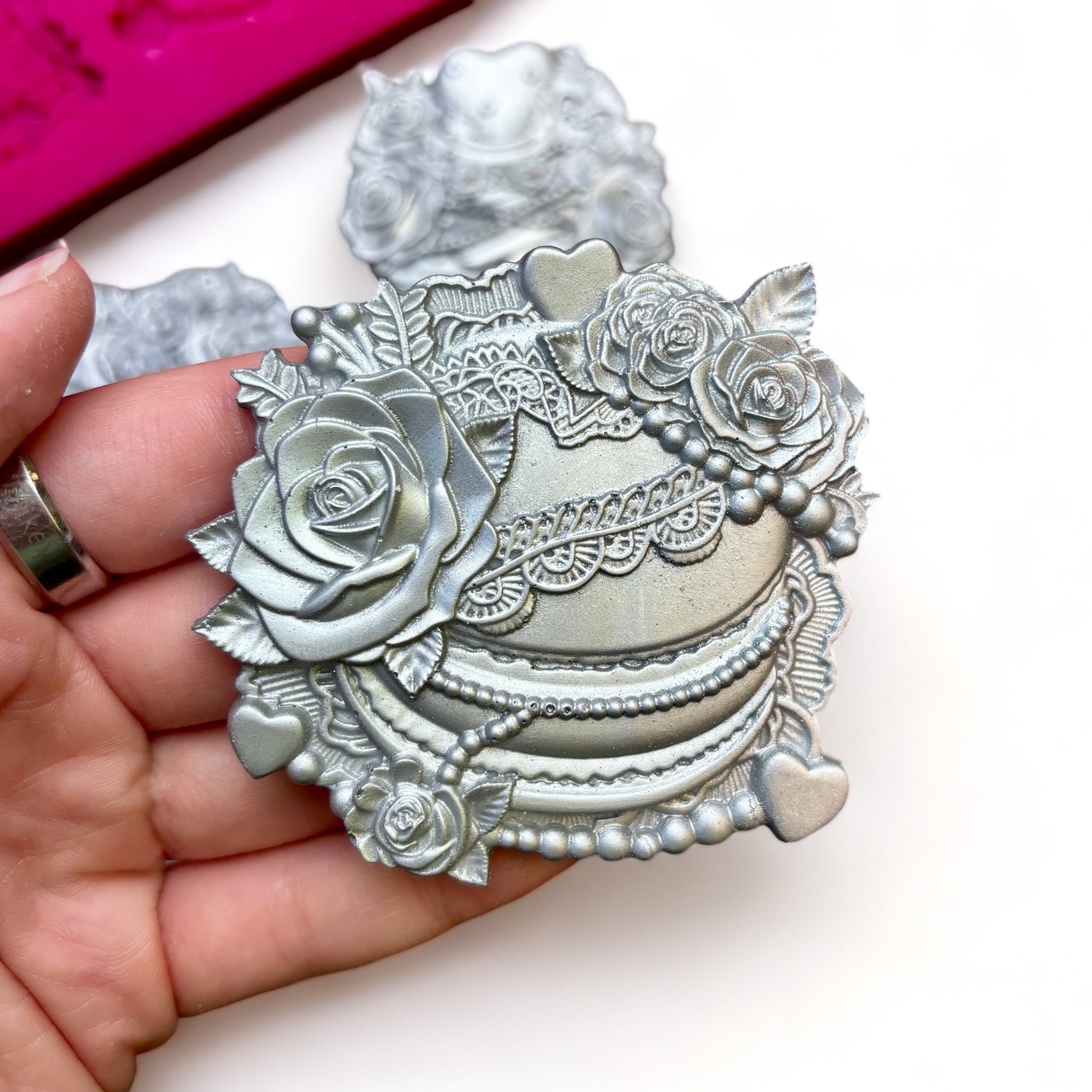 A pink silicone mold and silver colored castings of 4 Valentine's Day candy boxes surrounded with flowers are against a white background. A hand is shown holding one of the castings.