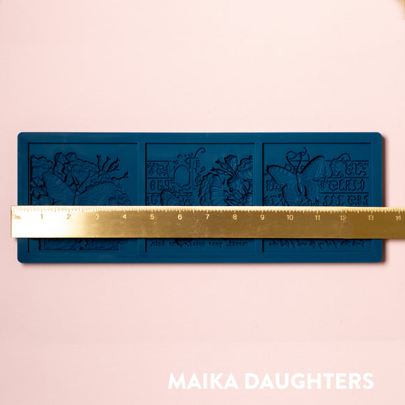 A blue silicone mold of 3 tiles featuring butterflies, flowers and calligraphy is against a light pink background. A gold ruler reading 13.4" length sits on the mold.