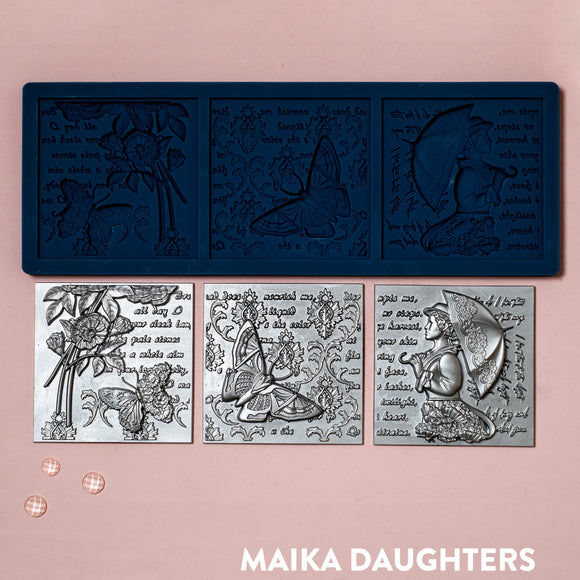 A blue silicone mold and silver colored castings of 3 tiles featuring a woman holding an umbrella, butterflies, flowers, and script writing are against a light pink background.