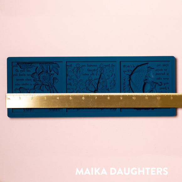 A blue silicone mold of 3 tiles featuring a woman holding an umbrella, butterflies, flowers, and script writing is against a light pink background. A gold ruler reading 13.4" length sits on the mold.