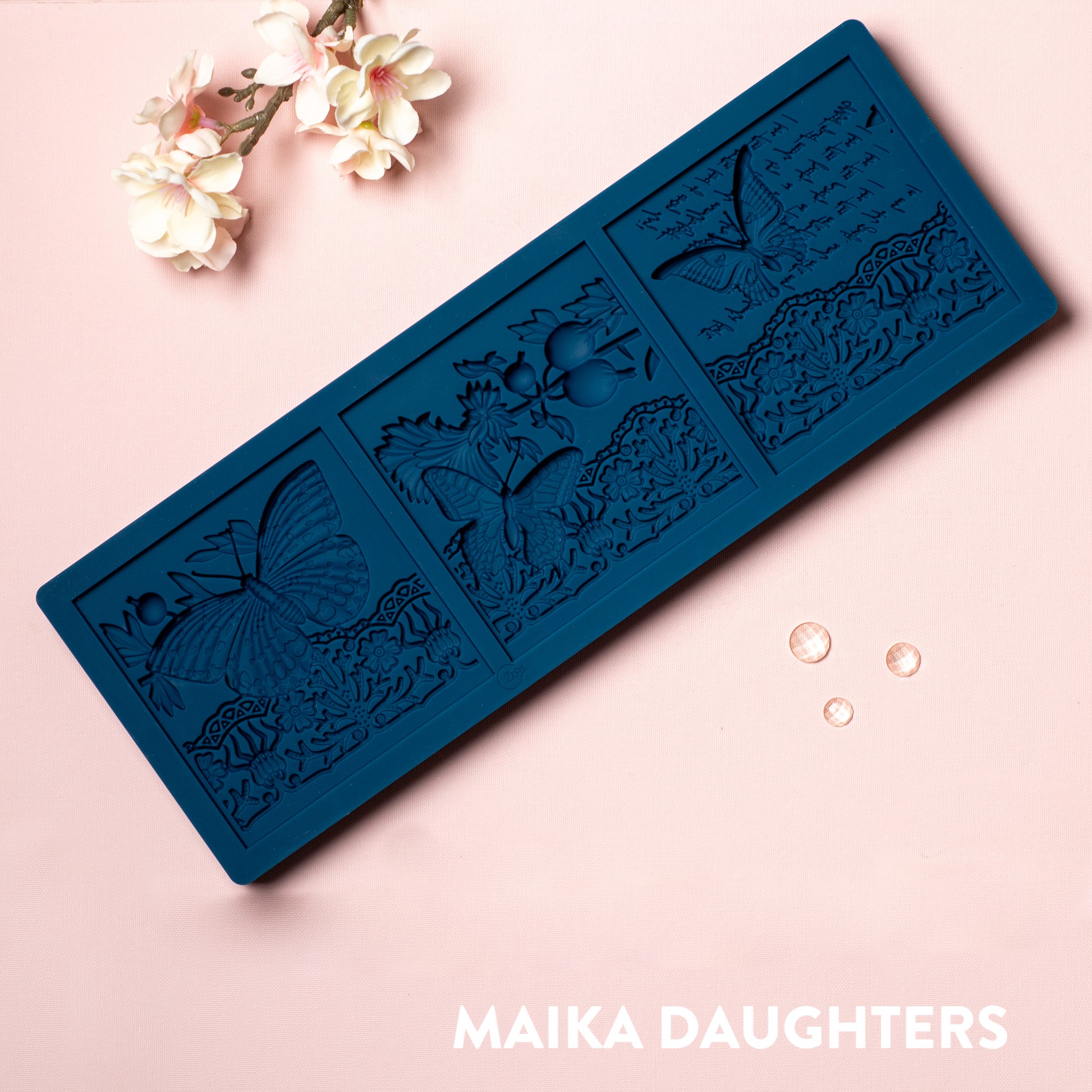 A blue silicone mold of 3 tiles featuring butterflies, a lace border, and script writing is against a light pink background.