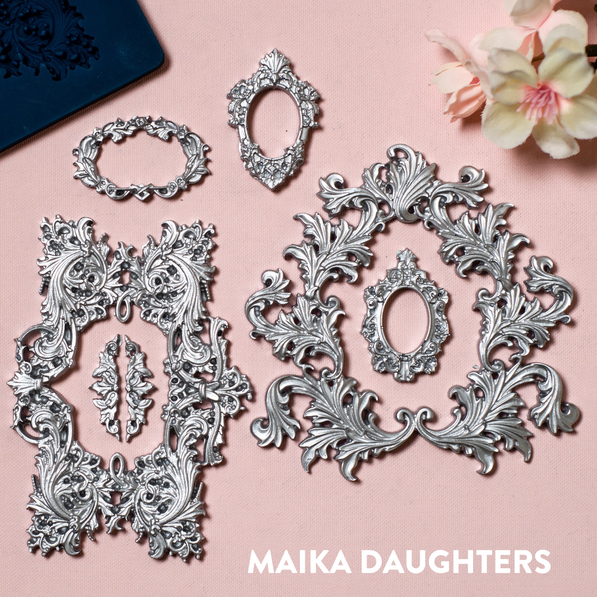 A blue silicone mold and silver colored castings of varying size ornate frames are against a light pink background.