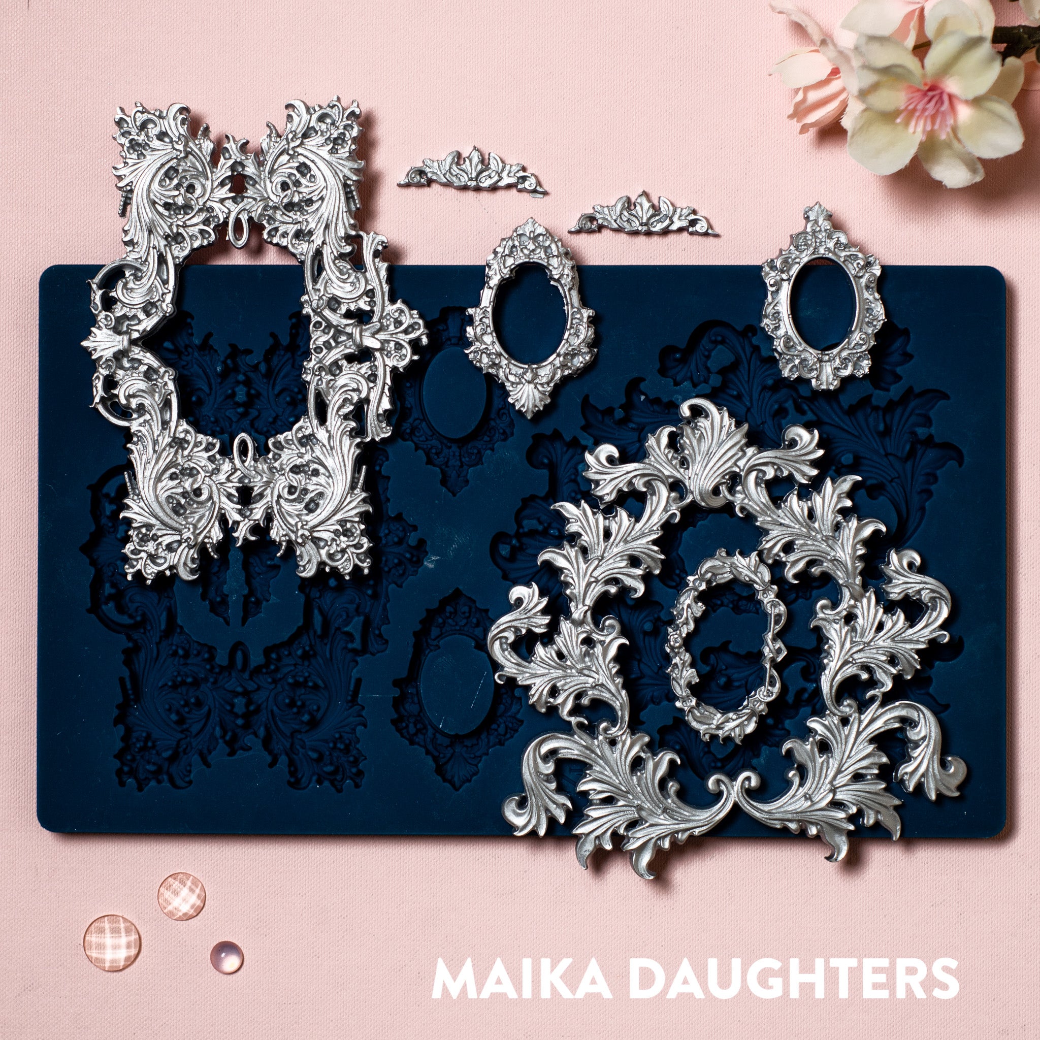 A blue silicone mold and silver colored castings of Zuri Design's Baroque Frames are against a light pink background.