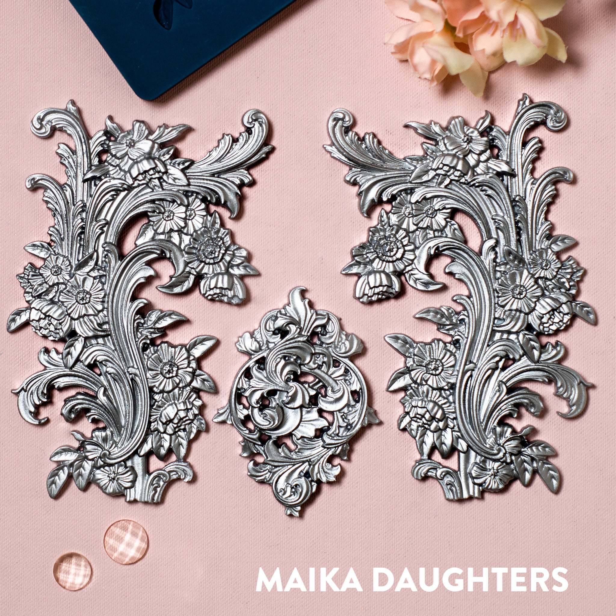 A blue silicone mold and silver colored castings of 2 large ornate floral flourishes and a small accent center piece are against a light pink background.