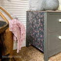 Close-up of a vintage 2-drawer nightstand refurbished by Annie Loveday Furniture is painted a grey-toned sage green and features the Peony Pattern A1 rice paper on the side. To the left is a wicker basket with a light pink blanket hanging out.