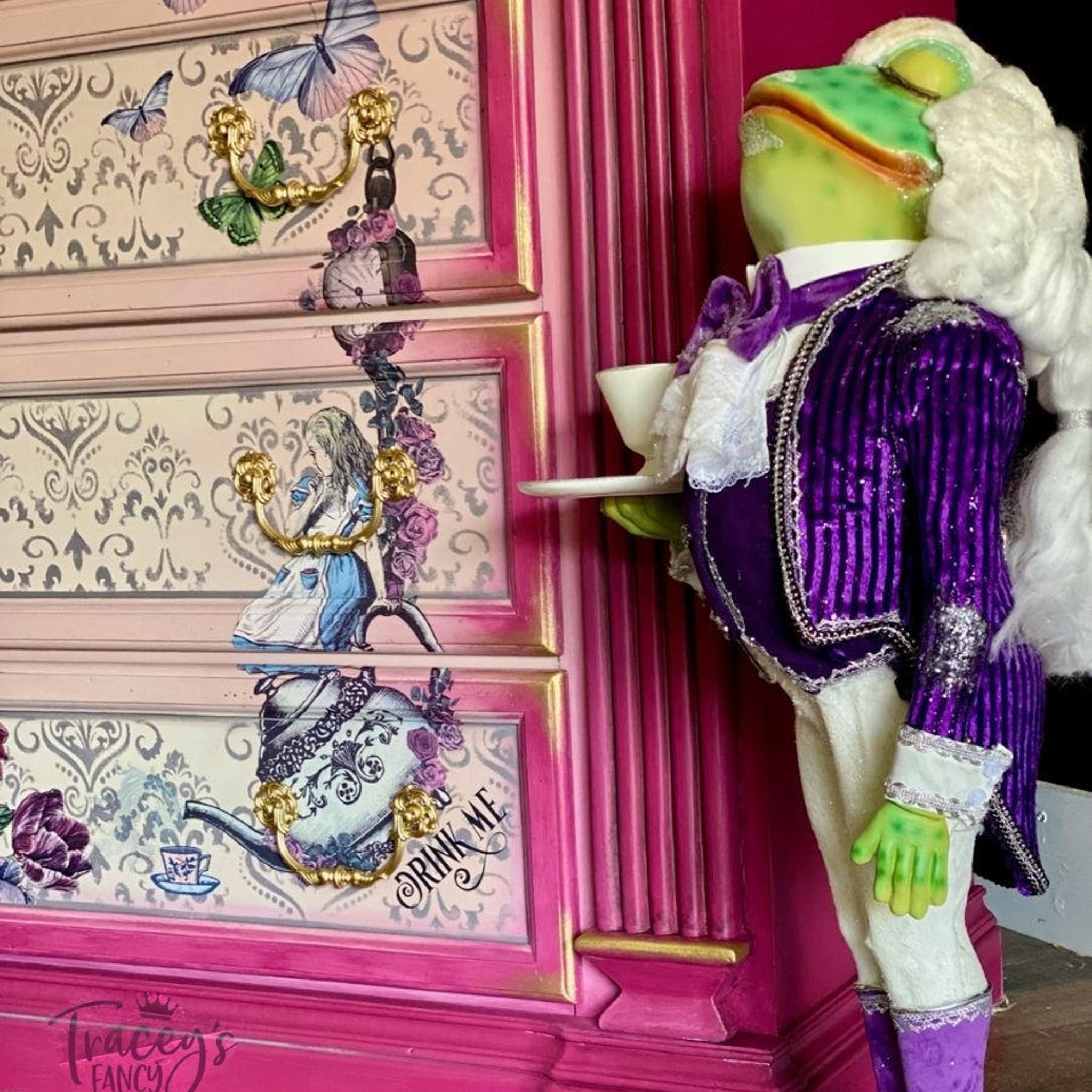 A close-up of a dresser refurbished by Trancey's Fancy is painted light pink and dark pink with gold accents features parts of the Alice Part 2 transfer. A statue of a frog waiter in a white wig and purple coat and vest stands next to the dresser.