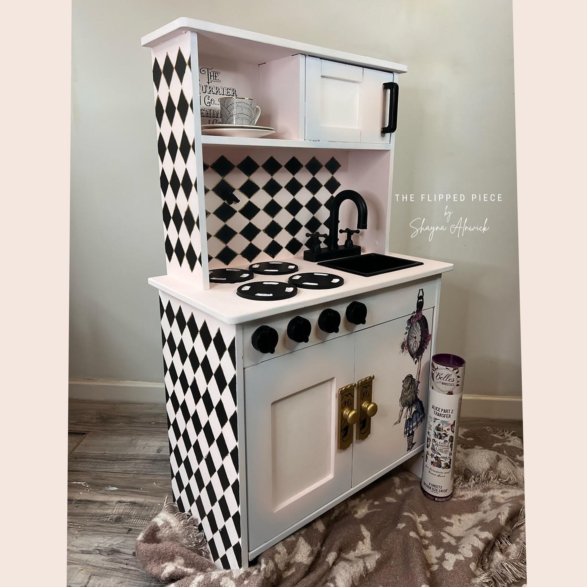A children's play kitchen refurbished by The Flipped Piece by Shayna Alnwick has been painted white and pale pink with black harlequin diamonds and features parts of the Alice Part 2 transfer.
