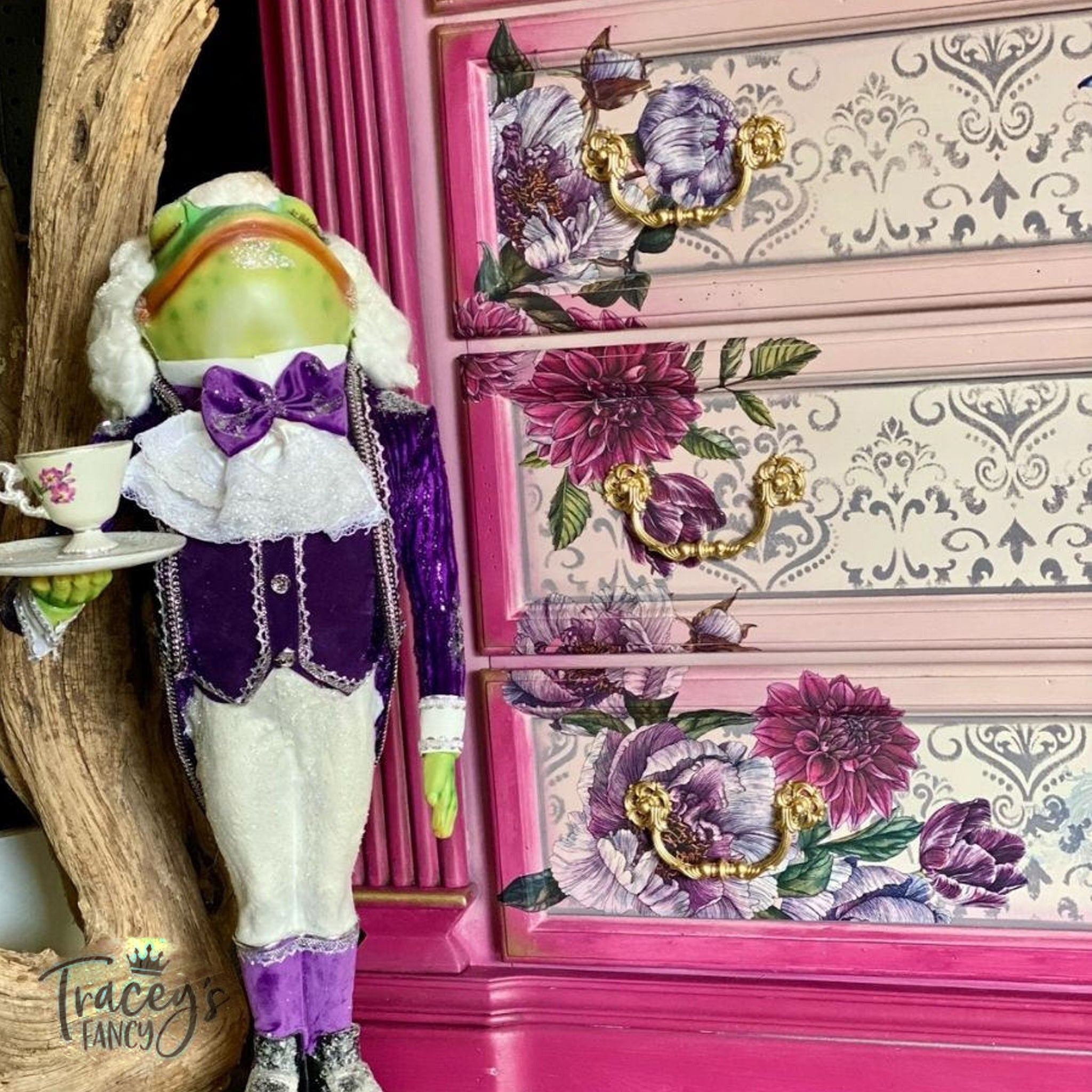 A close-up of a dresser refurbished by Trancey's Fancy is painted light and dark pink with gold accents and features the Buds and Branches transfer. A statue of a frog waiter in a white wig and purple coat and vest stands next to the dresser.
