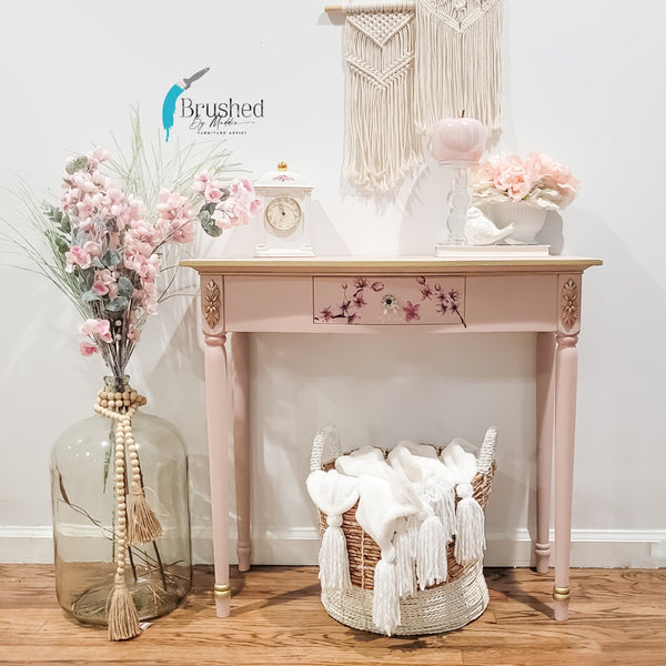 A pale pink side table refurbished by Brushed by Maddie Furniture Artist features the Cherry Blossom transfer on a small drawer. A large glass jug with flowers sits to the left of the table. A whicker basket with white cloth sits under the table and decorations sit on top.