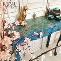 A skinny side table refurbished by Revu & Corrigé is painted a sky blue and features the Cherry Blossom transfer. A tall cherry blossom plant sits to the left of the table and a gold cat statue and black teapot set are on the table.