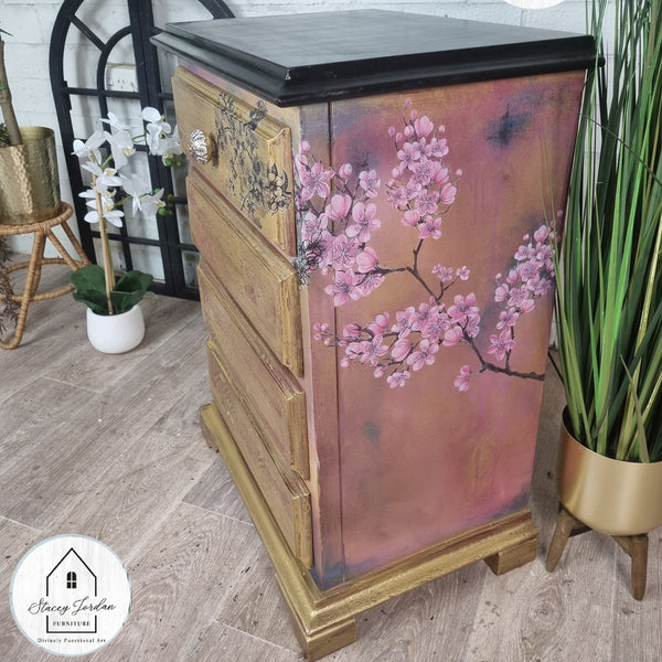 A vintage 4-drawer nightstand refurbished by Stacey Jordan Furniture Divinely Functional Arts is painted a blend of coral pink and gold with a black top and features the Cherry Blossom transfer on its sides.