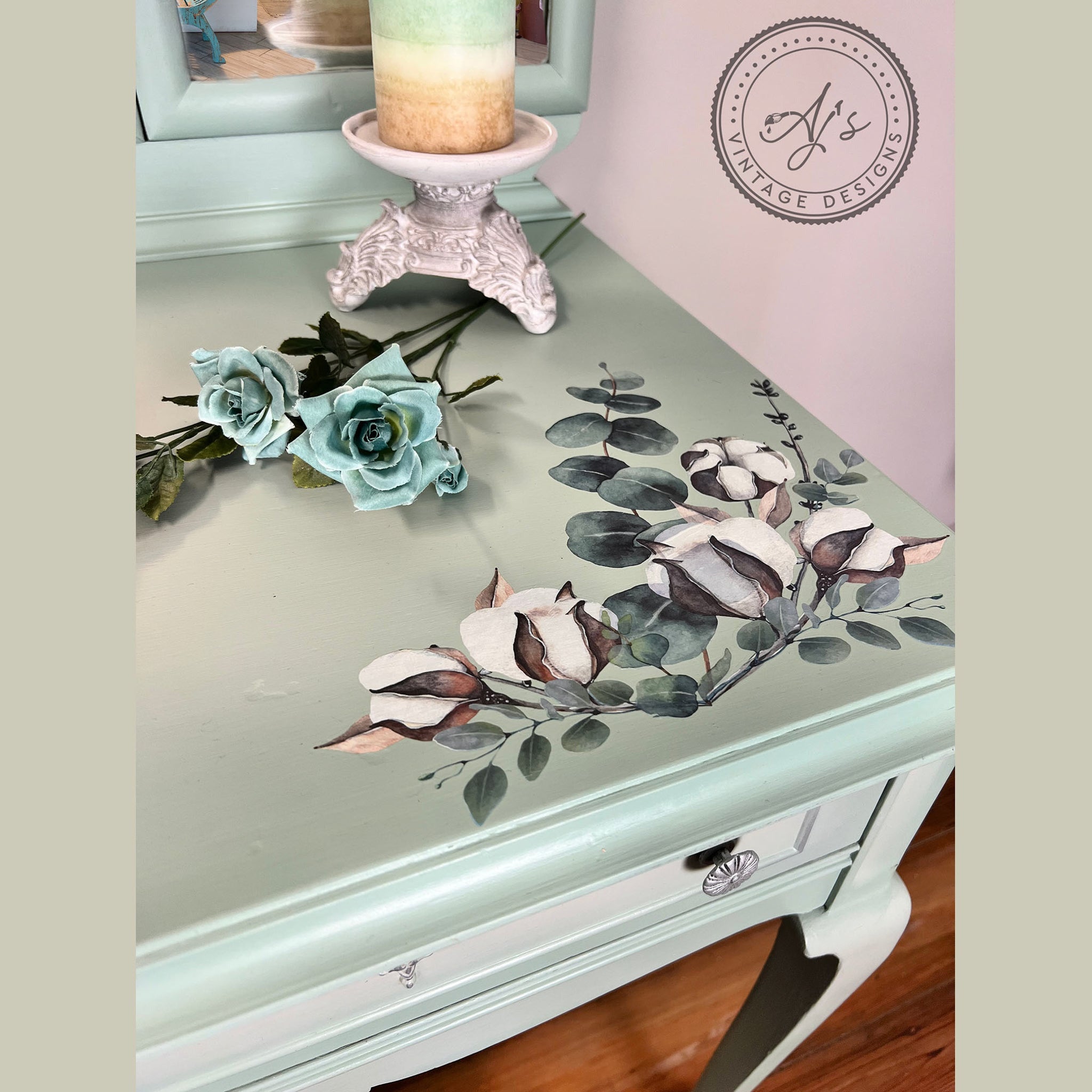 A close-up of a vintage vanity desk and sitting stool refurbished by Aj's Vintage Designs is painted sea foam green and features the Cotton and Eucalyptus transfer on the top of the vanity.