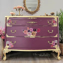 A vintage 3-drawer dresser refurbished by Chic and Shabby Furniture by Rebecca is painted a plum purple with gold accents and features the Flower Child transfer on its drawers.
