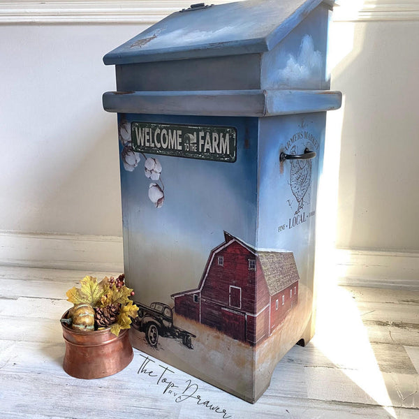A vintage wood trash bin refurbished by The Top Drawer is painted a sky blue with clouds and dusty prairie brown and features the large red barn, truck and other designs from the On the Farm transfer.