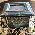 A vintage jewelry box refurbished by TLC Vintage is painted black with patina accents and features the Retro Peacock trasnfer. Costume pearl necklaces and white flowers sit around the front of the box.
