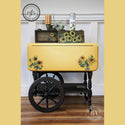 A vintage bar cart refurbished by AJ's Vintage Designs, a Dixie Belle Paint Company Brand Ambassador, is painted black with a yellow collapsible top. On the cart are 2 small wood projects and they all feature the Sunflowers Transfer on them.