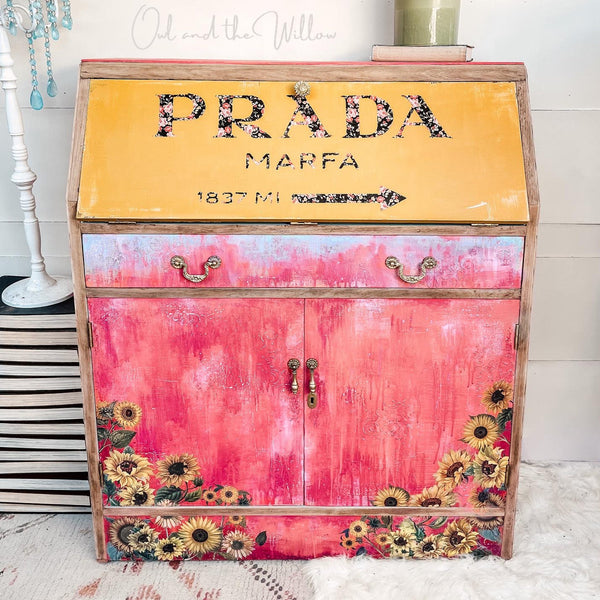 A vintage secretary desk is painted a blend of red and light orange on the front and features the Sunflowers Transfer. The pull-down top of the desk is painted yellow and has text that reads: Prada Marfa 1837 Miles with an arrow pointing to the right.