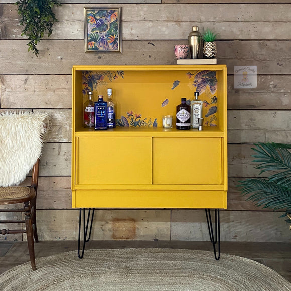 A mid-century bar cabinet refurbished by Cherbs Chalks Interiors is painted in Dixie Belle's Colonel Mustard chalk mineral paint.