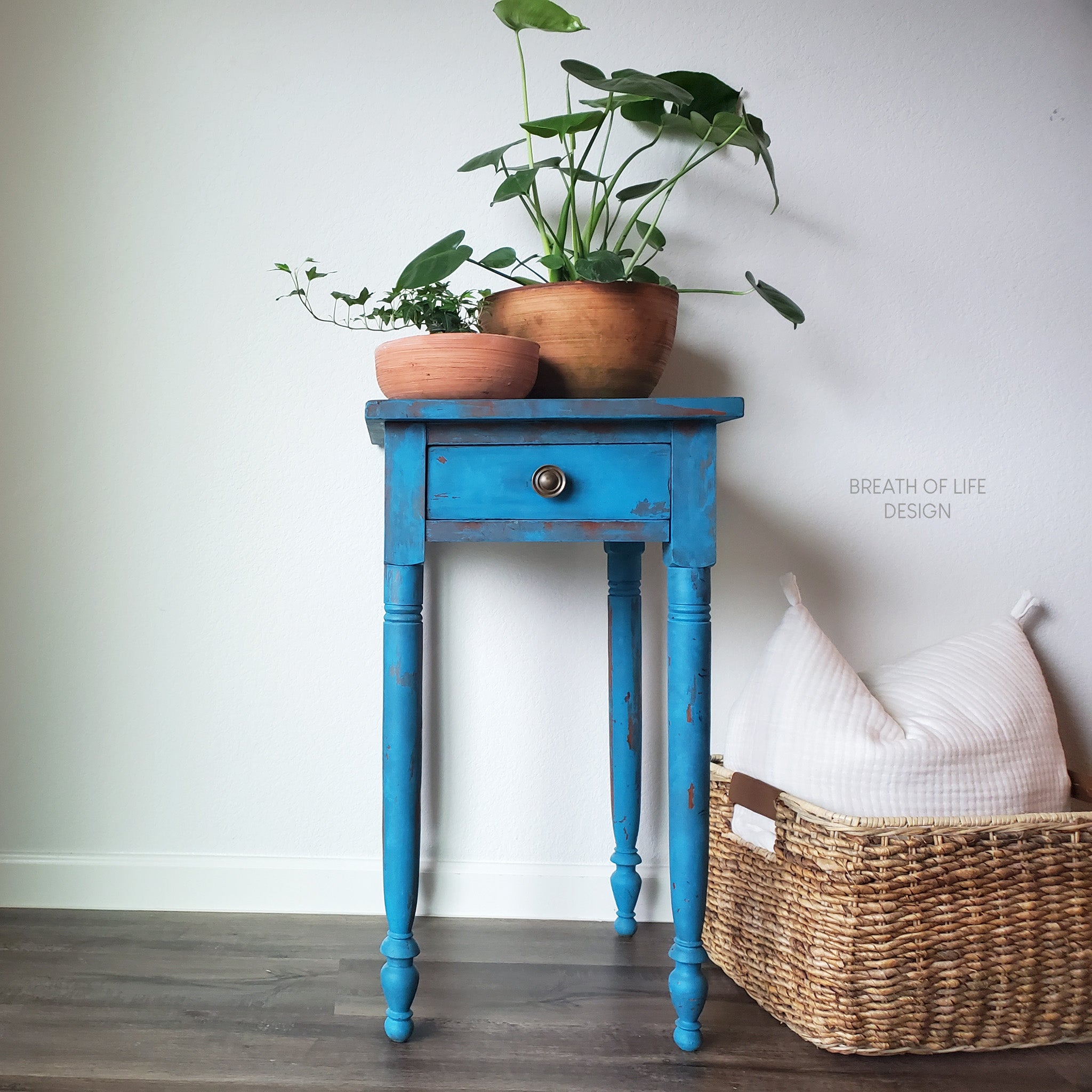 A small vintage side table refurbished by Breath of Life Design is painted in Peacock Chalk Paint. Two plant pots sit on the table and a wicker backet with a white pillow sits on the floor to the right of the table.