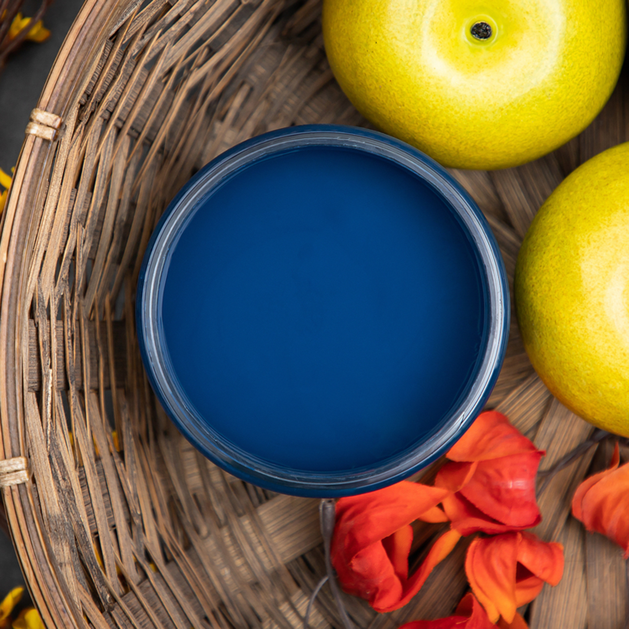 An arial view of an open container of Dixie Belle Paint Company's Bunker Hill Blue Chalk Mineral Paint is in a wicker basket surrounded by fruit and flowers.