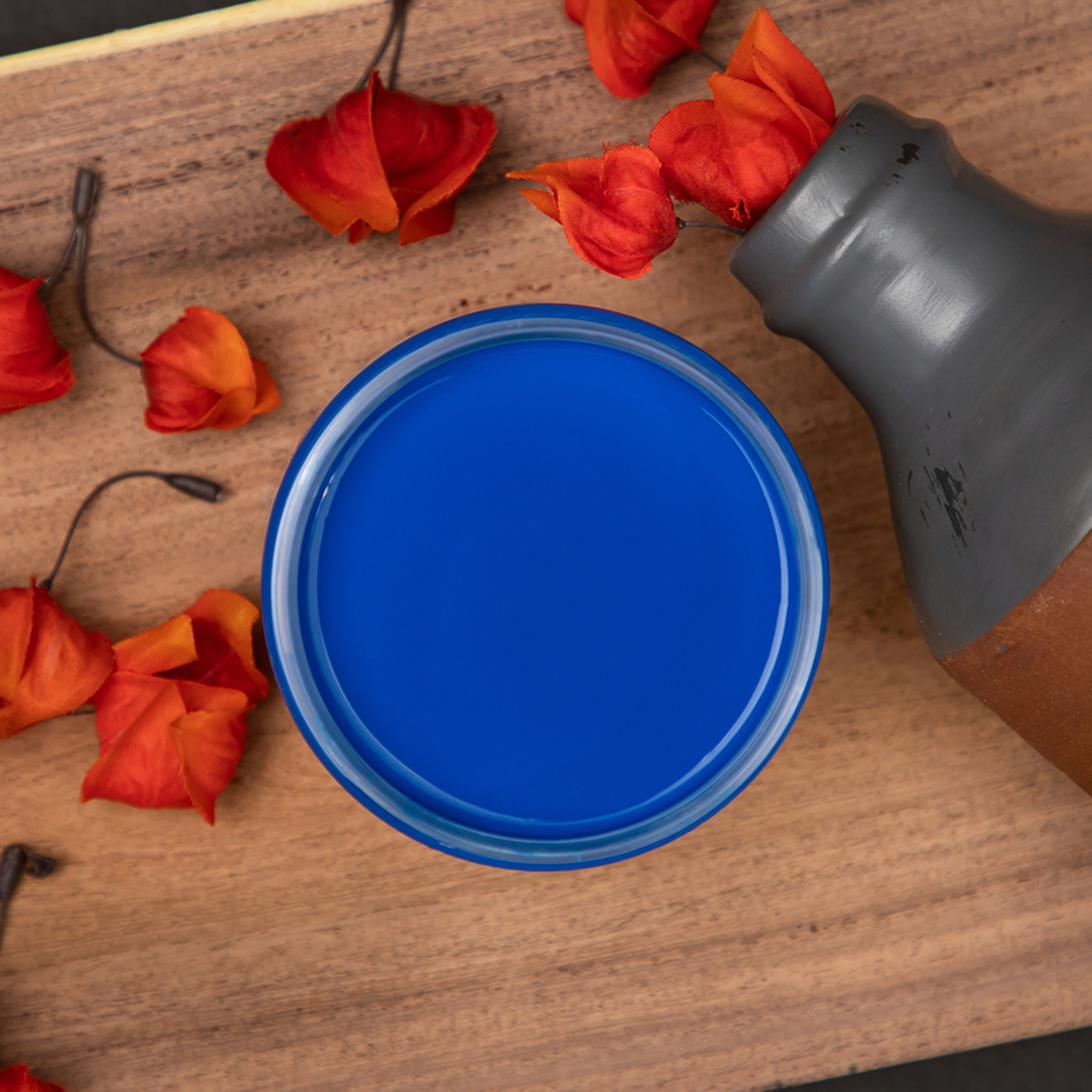 An arial view of an open container of Dixie Belle Paint Company's Cobalt Blue Chalk Mineral Paint is surrounded by red-orange silk flowers coming out of a silver painted bottle against a wood background.