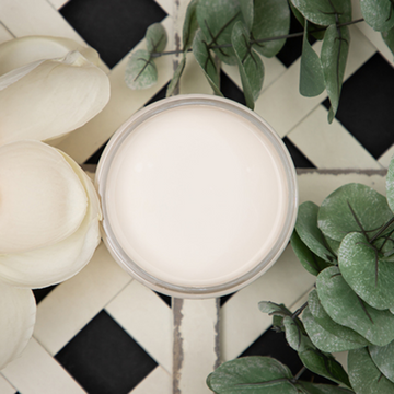 An arial view of an open container of Dixie Belle Paint Company’s Drop Cloth Chalk Mineral Paint is surrounded by silk green leaves and cream-colored silk flowers against lattice painted in Drop Cloth.
