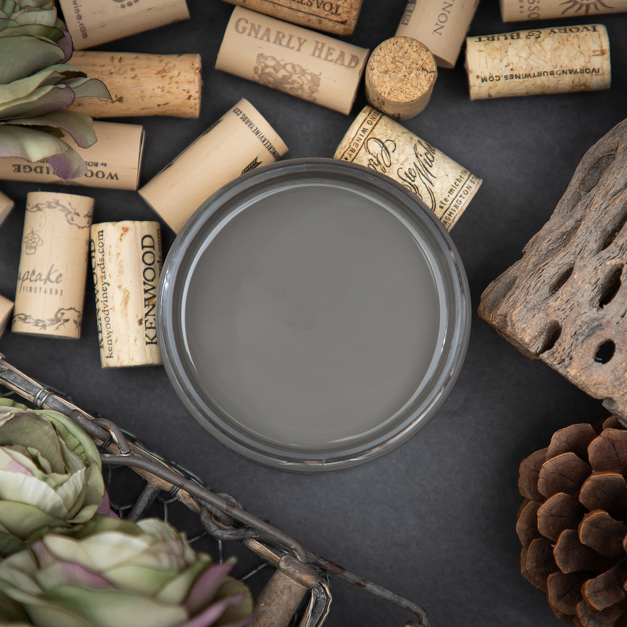 An arial view of an open container of Dixie Belle Paint Company’s Gravel Road Chalk Mineral Paint is surrounded by cork toppers, flowers, and pine cones.