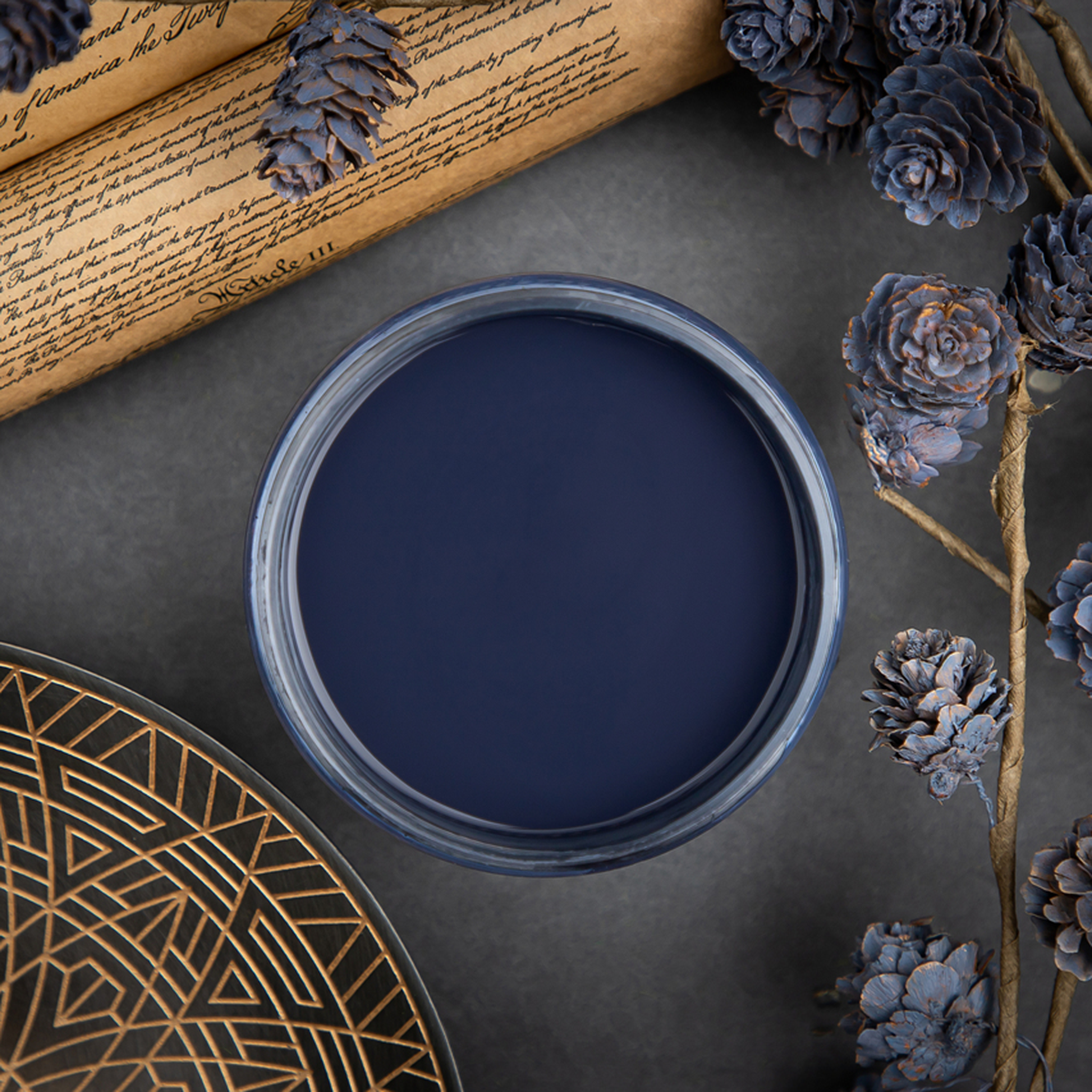 An arial view of an open container of Dixie Belle Paint Company’s In the Navy Chalk Mineral Paint is surrounded by blue painted pine cones and an old parchment scroll.