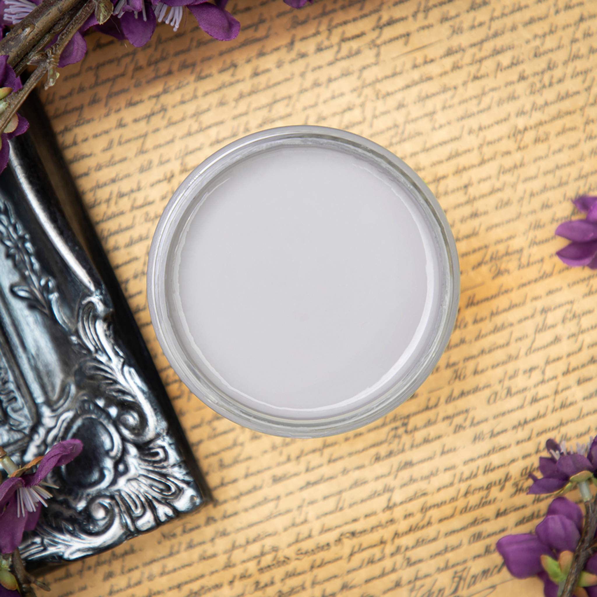 An arial view of an open container of Dixie Belle Paint Company’s Mason Dixon Chalk Mineral Paint is sitting on old parchment with script on it and surrounded by small purple silk flowers and a silver photo frame.