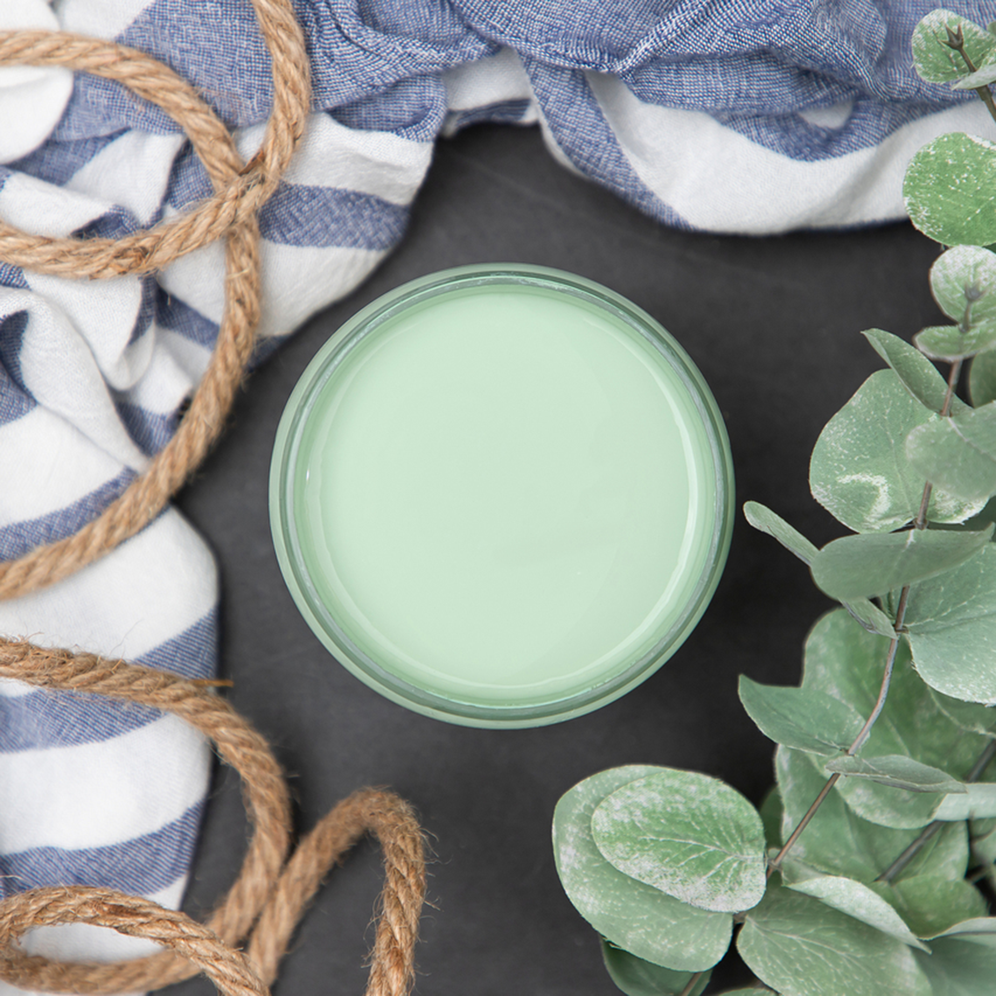 An arial view of an open container of Dixie Belle Paint Company’s Mint Julep Chalk Mineral Paint is surrounded by eucalyptus, rope, and a blue and white striped piece of linen.