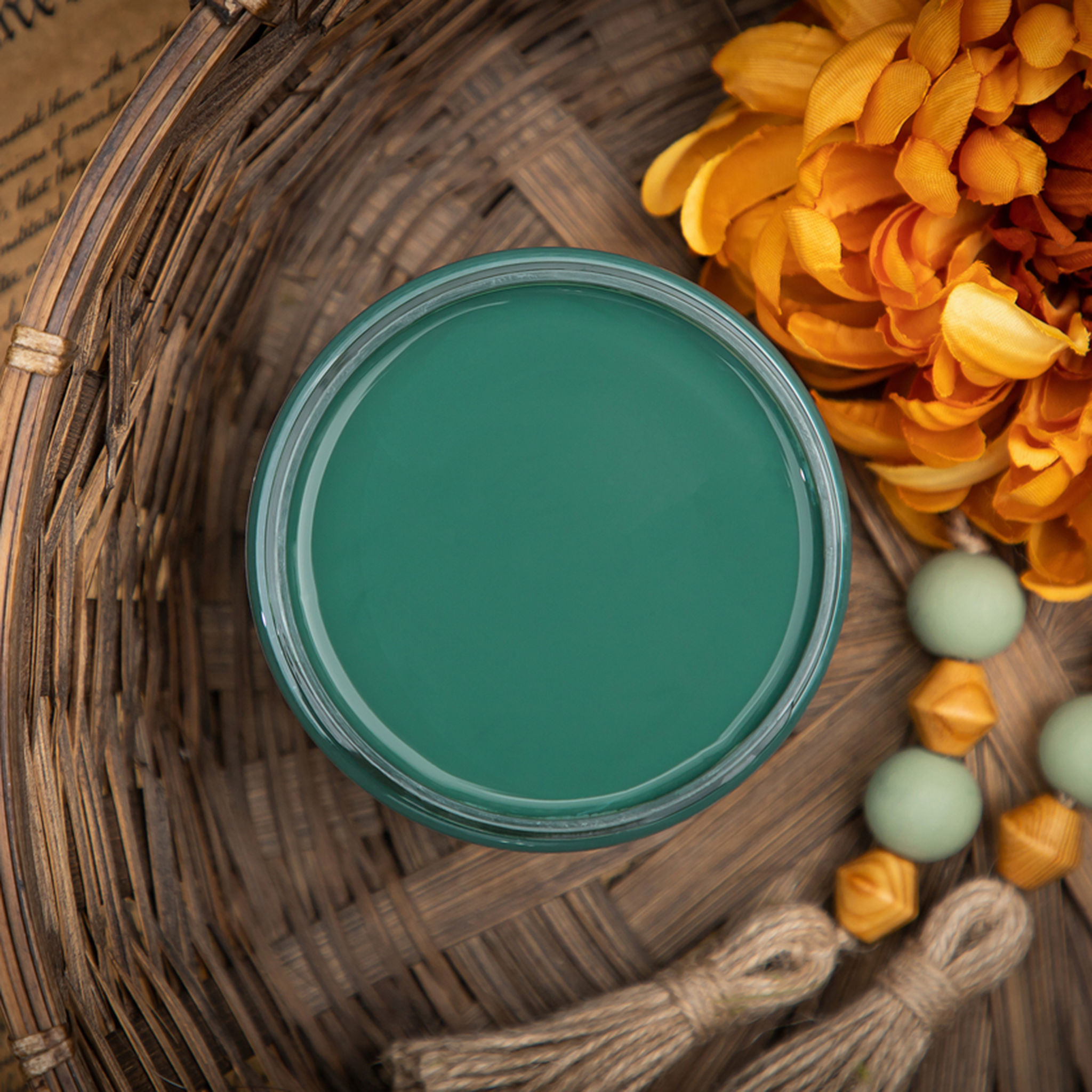 An arial view of an open container of Dixie Belle Paint Company’s Palmetto Chalk Mineral Paint is in a wicker basket with an large orange silk flower with wood beads and twine tassles.