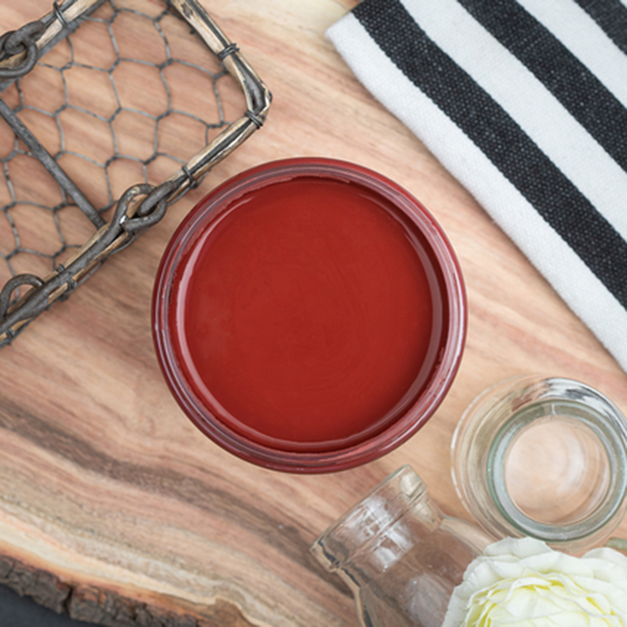 An arial view of an open container of Dixie Belle Paint Company’s Rustic Red Chalk Mineral Paint is on a wood board and surrounded by black and white striped fabric and glass bottles.