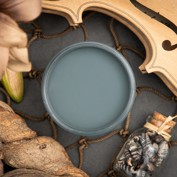 An arial view of an open container of Dixie Belle Paint Company’s Stormy Seas Chalk Mineral Paint is surrounded by a rope net, driftwood, and seashells in a glass bottle.
