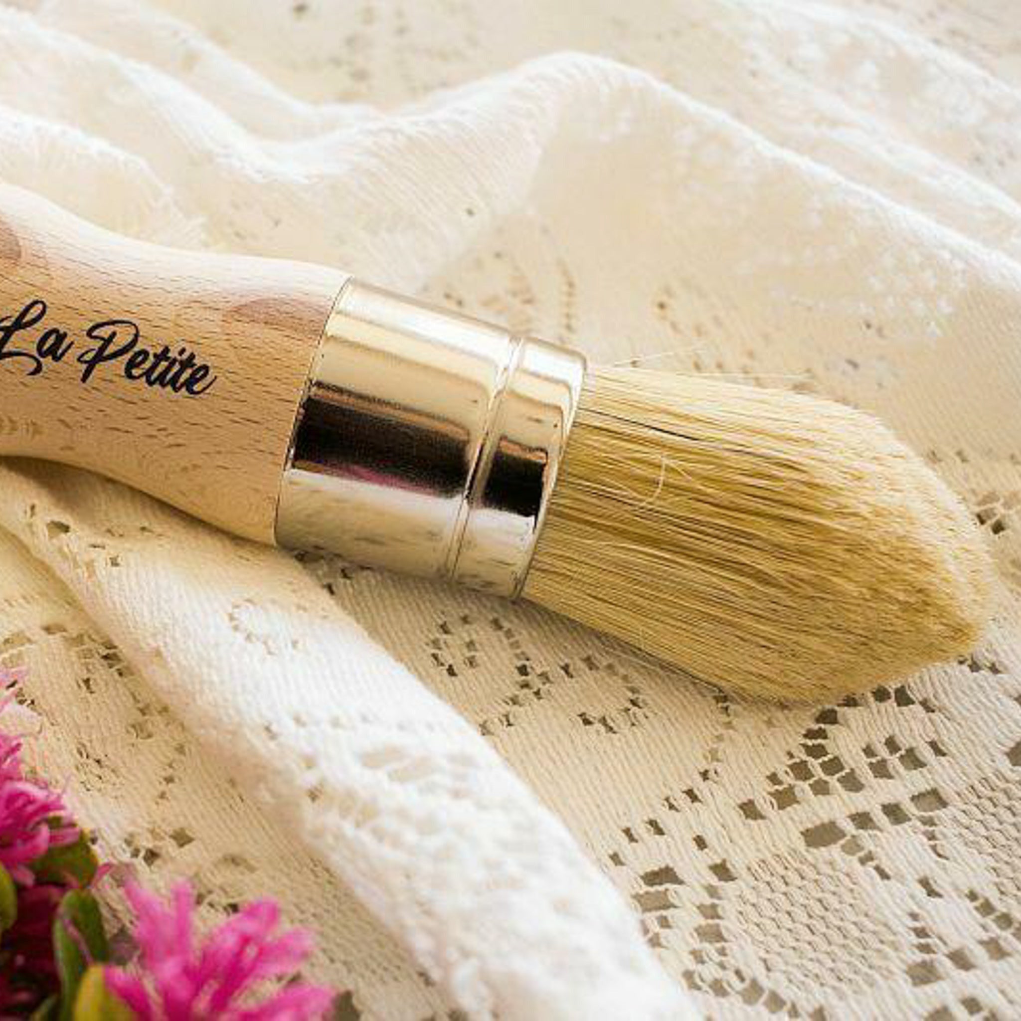 Dixie Belle's La Petite Paint Brush is against a lace background. This brush has natural bristles and is shaped to help reach the fine-detailed areas on your furniture projects.