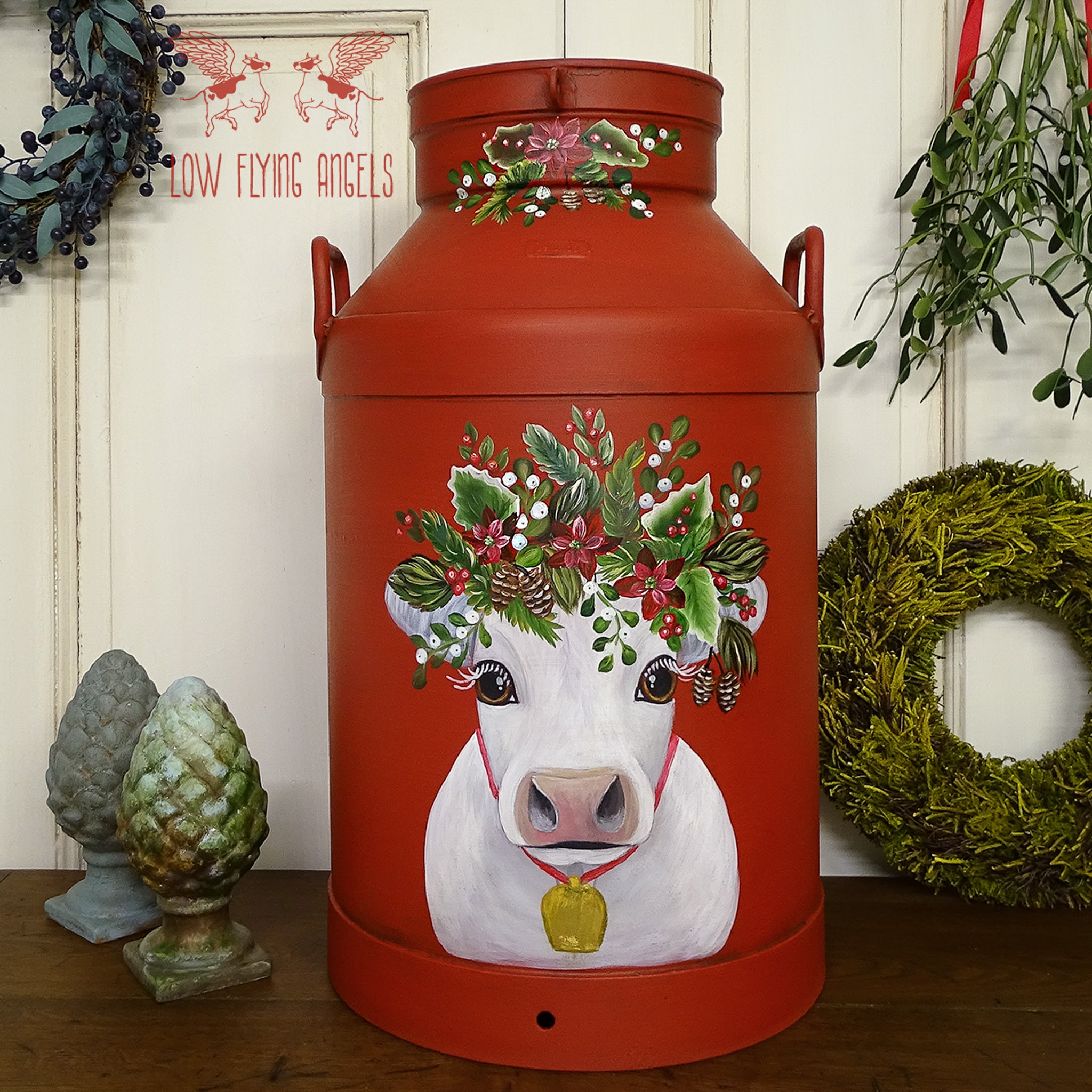 A vintage metal milk can refurbished by Low Flying Angels is painted with Dixie Belle's Barn Red chalk mineral paint and features a painting of a white cow wearing a Christmas wreath on its head.