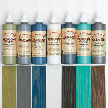 Seven 8oz containers of Dixie Belle's Voodoo Gel Stains are against a white background with their matching wood swatches under them. The colors are: Bayou Moss, Up In Smoke, Denim, White Magic, Black Magic, Temptress, and Tobacco Road.