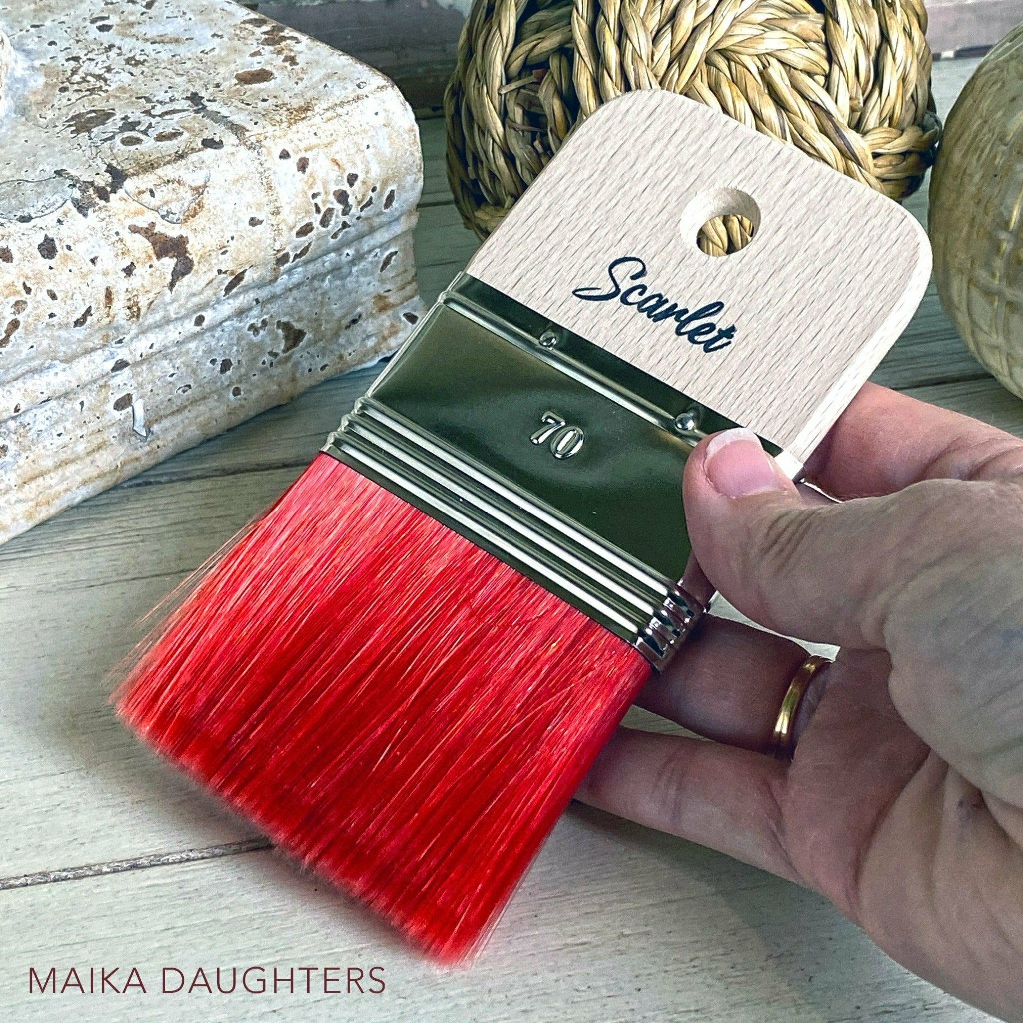 A hand is shown holding a wide rectangular shaped paint brush with scarlet red bristles against a wood table with  twine ball and a plaster piece of decoration.