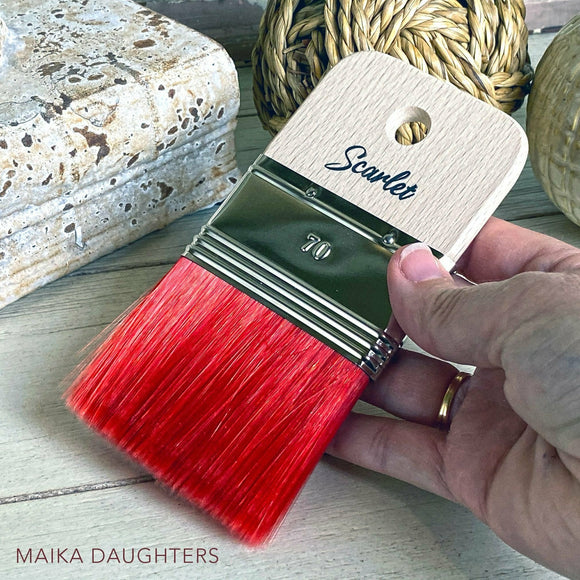 A hand is shown holding a wide rectangular shaped paint brush with scarlet red bristles against a wood table with  twine ball and a plaster piece of decoration.