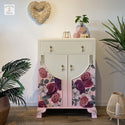 A small vintage dresser with 1 drawer and 2 cabinet doors refurbished by Cherub's Chalk Interiors is painted cream and light pink and features the Floral Romance transfer towards the bottom.