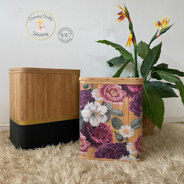 Two wood slated laundry hampers refurbished by Cherub's Chalks Interiors, a Dixie Belle Paint Company Content Creator, are natural wood stained. The larger hamper has black and gold on the bottom and the smaller hamper features the Floral Romance transfer. A large Bird of Paradise plant sits behind the hamper on the right.