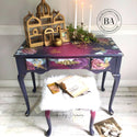 A vintage vanity desk and stool refurbished by The Top Drawer, a Dixie Belle Paint Company Brand Ambassador, is painted dark purple and pink and features the Floral Romance transfer on it. A gold India style bird cage and candles sit on top of the vanity.