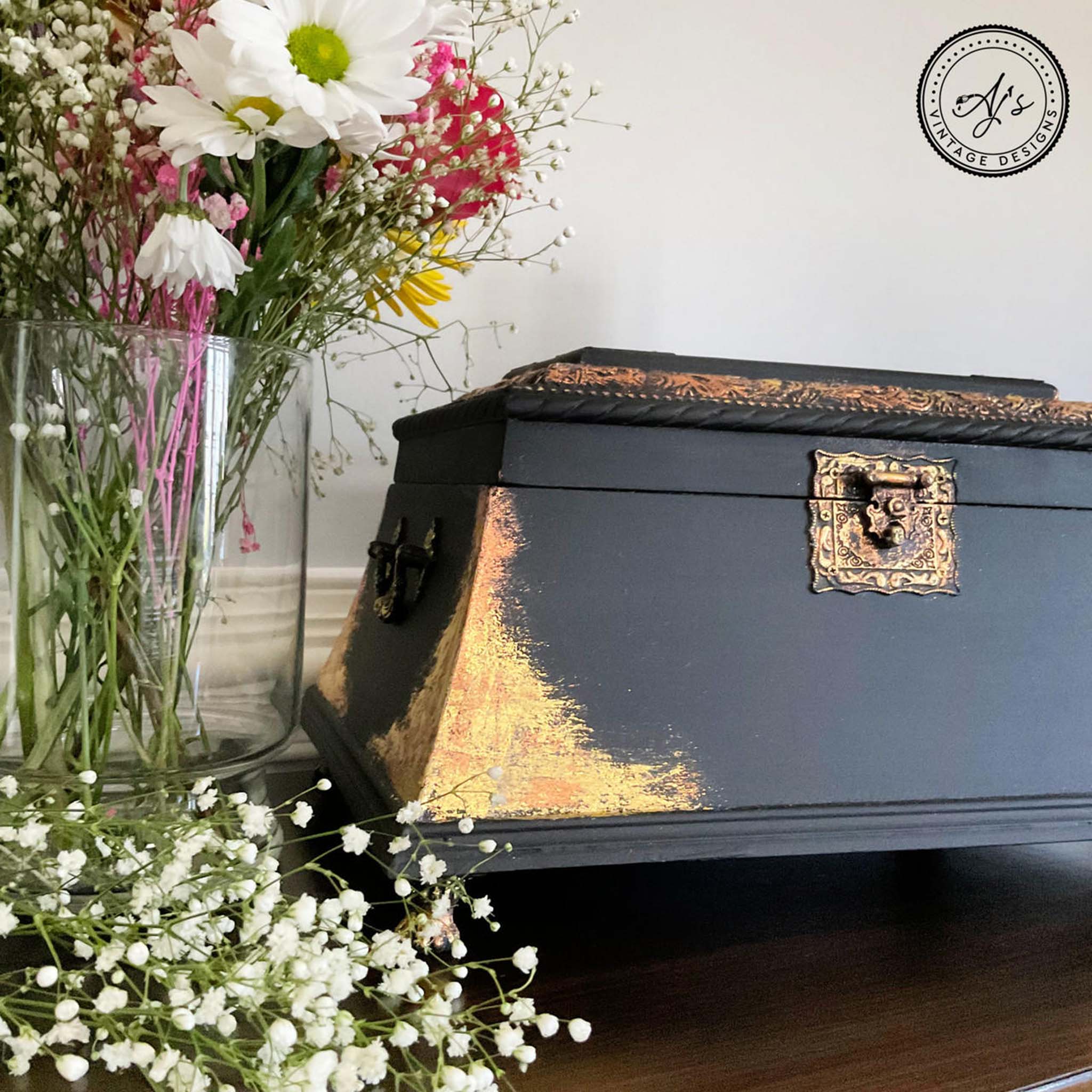 A small vintage box refurbished by Aj's Vintage Designs is painted black with Dixie Shine gold foil on the corners. To the left is a glass vase with small wildflowers in it.