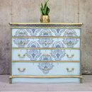 A 4-drawer dresser refurbished by Kacha is painted light blue with gold accents and features the Kacha Dana Damask transfer on it.