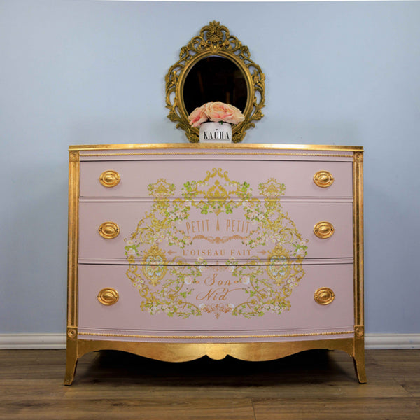 A vintage 3-drawer dresser refurbished by Kacha is painted a soft pink with gold accents and features the Kacha Petite A Petite transfer on its drawers.