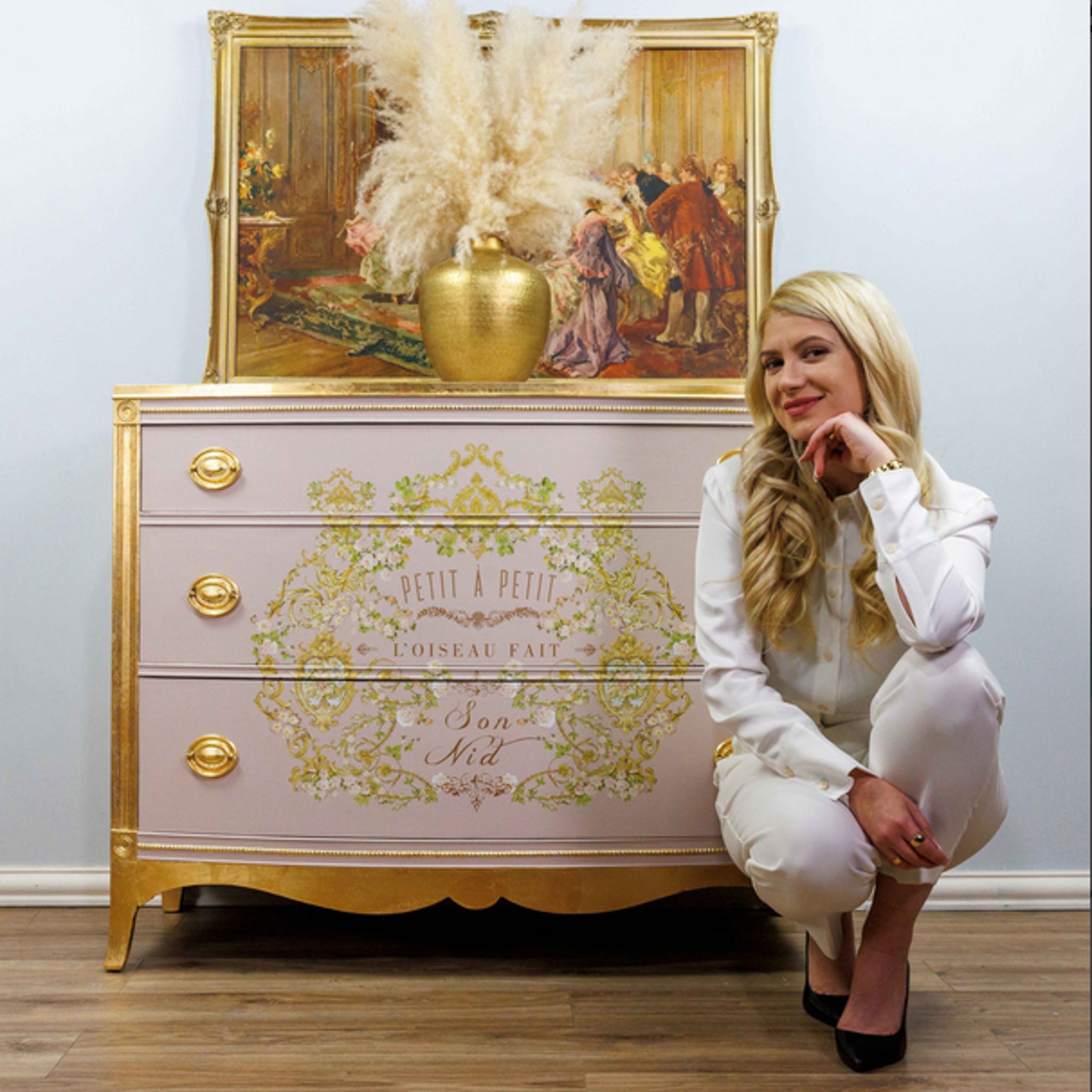 A vintage 3-drawer dresser is painted a soft pink with gold accents and features the Kacha Petite A Petite transfer on its drawers. The designer Kacha is knelt down in front of of the right side of the dresser.