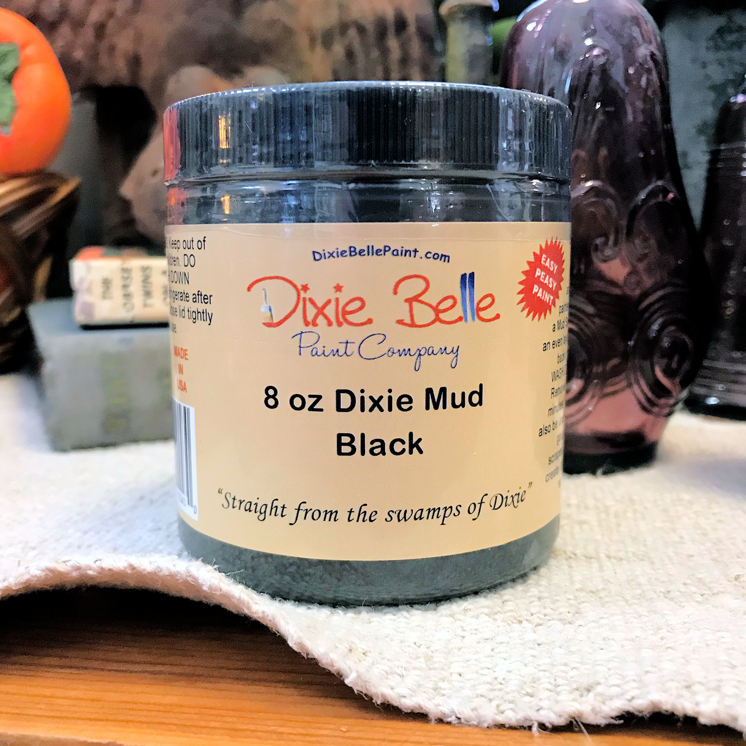 An 8oz container of Dixie Mud by Dixie Belle Paint Company in Black. Sitting on a cream-color table cloth with a basket and a purple glass item in the background.