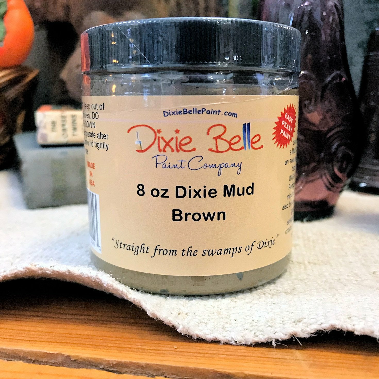 An 8oz container of Dixie Mud by Dixie Belle Paint Company in Brown. Sitting on a cream-color table cloth with a basket and a purple glass item in the background.