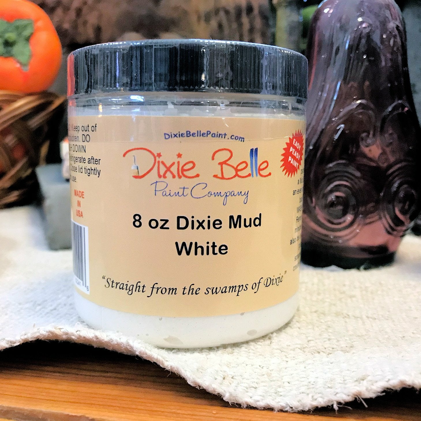 An 8oz container of Dixie Mud by Dixie Belle Paint Company in White. Sitting on a cream-color table cloth with a basket and a purple glass item in the background.