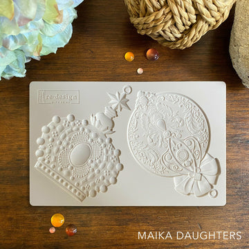 Wooden table with a mold of two ornaments. A white Maika Daughters logo is in the bottom right corner.