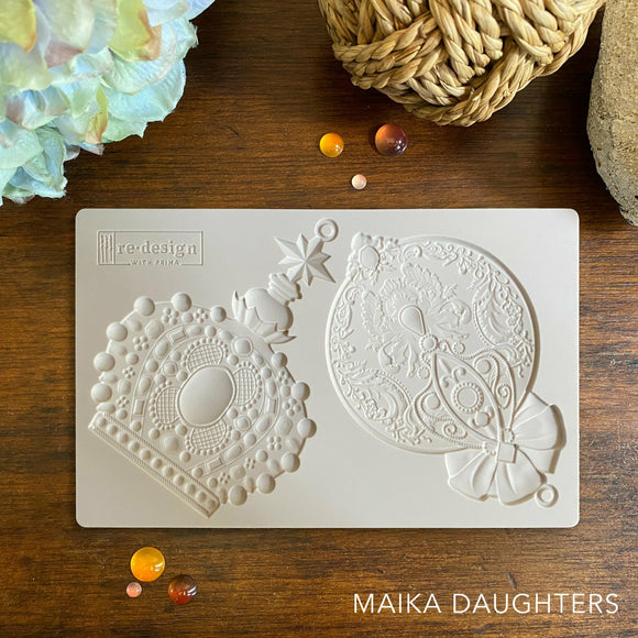 Wooden table with a mold of two ornaments. A white Maika Daughters logo is in the bottom right corner.